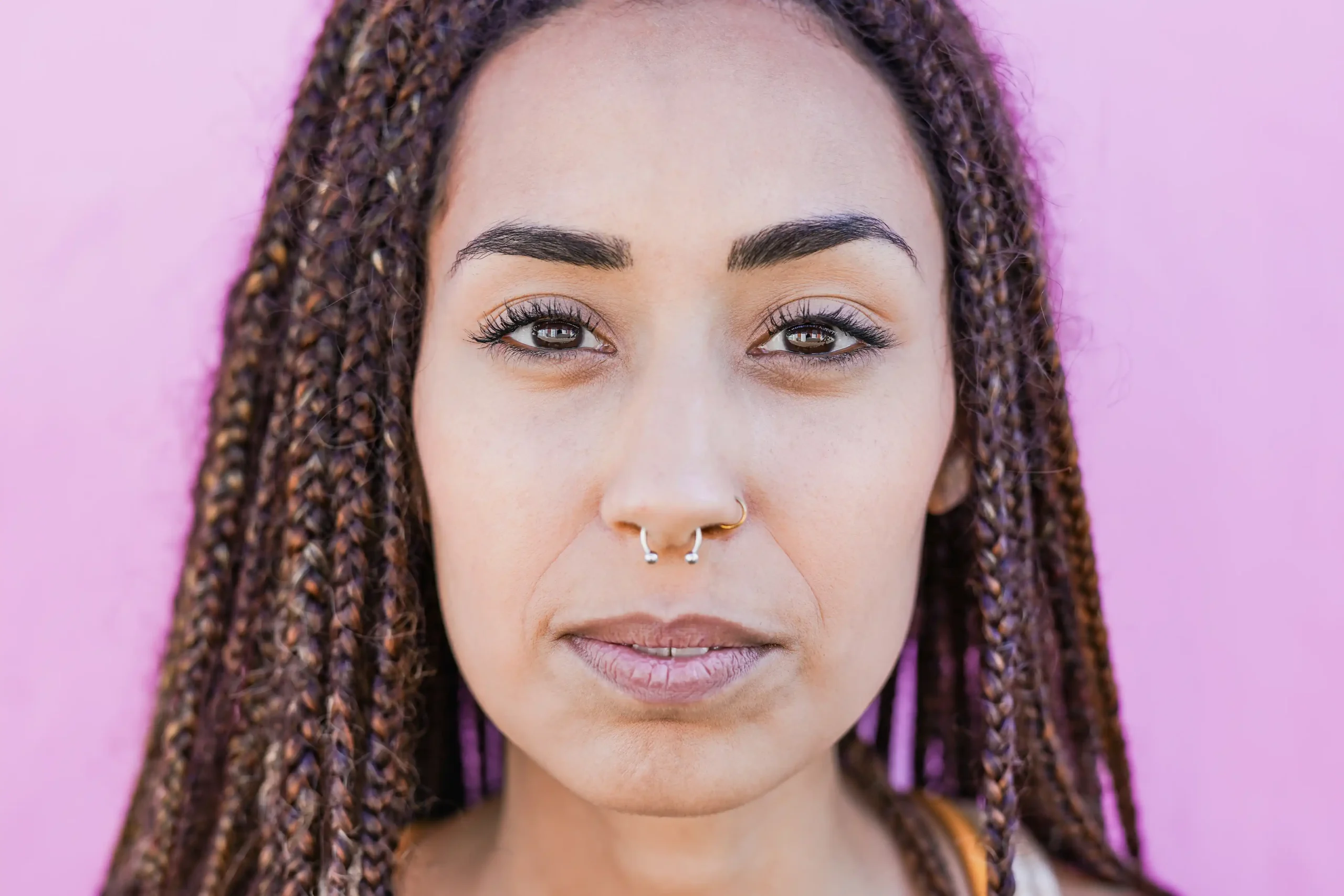 A imagem mostra uma jovem mulher, com expressão séria, com tranças no cabelo, piercing no nariz, olhando para a câmera.