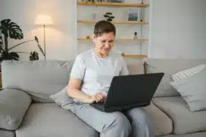 A imagem mostra uma mulher de meia idade, sorridente, usando o laptop, enquanto está sentada em um sofá, dentro de casa. Ilustra o texto sobre Renovação da Carteira de Registro Nacional Migratório.