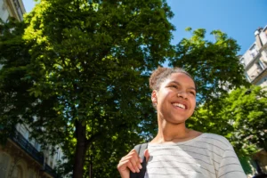 A imagem mostra uma jovem negra, sorrindo, com uma mochila nas costas, andando pela cidade. Ilustra o texto sobre refugiados.