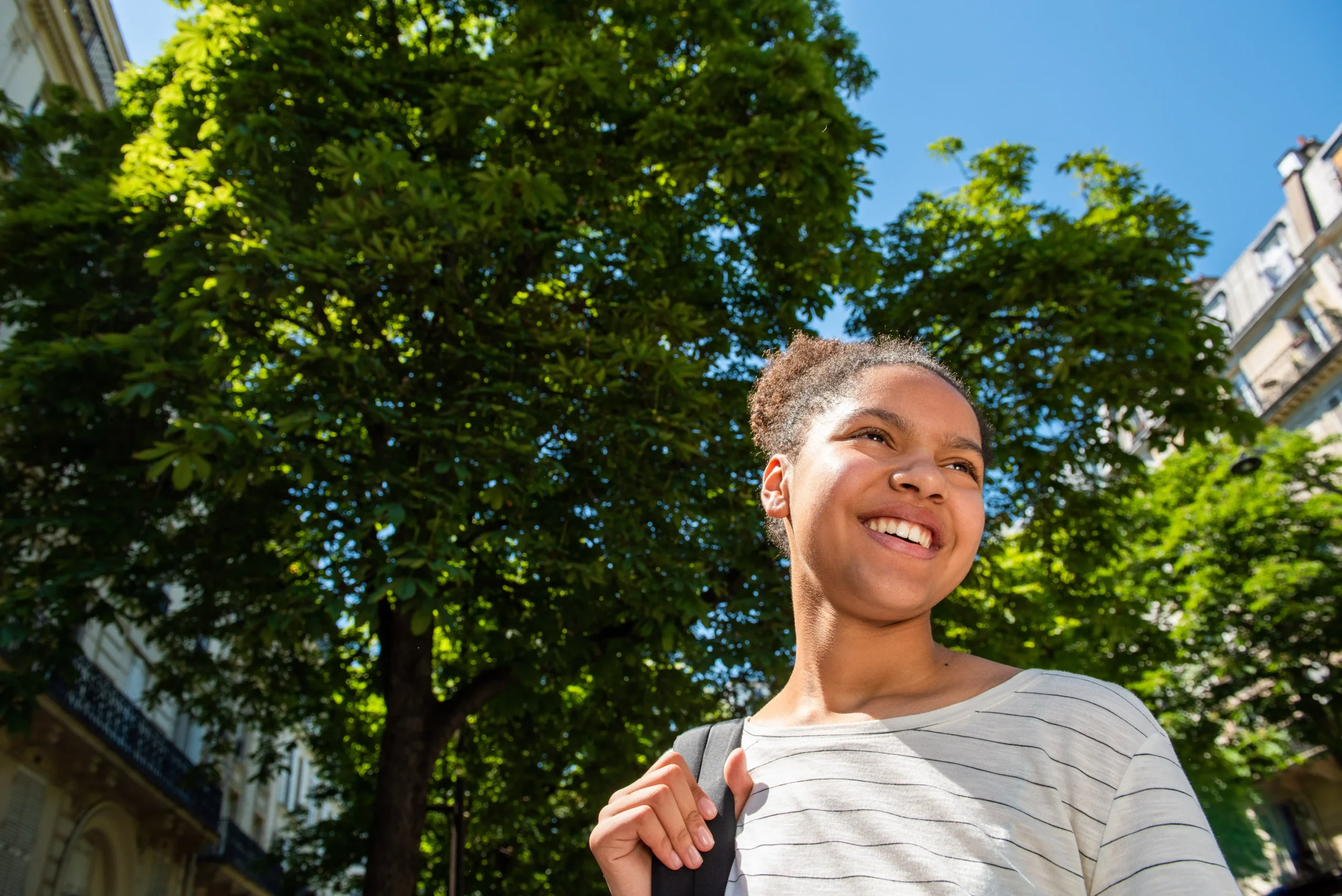 A imagem mostra uma jovem negra, sorrindo, com uma mochila nas costas, andando pela cidade. Ilustra o texto sobre refugiados.