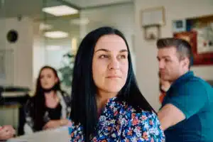 A imagem mostra uma mulher de meia idade, com cabelos longos e pretos, olhando para o horizonte em meio a uma reunião com outras 2 pessoas. Ilustra o texto sobre serviço de apoio a refugiados.