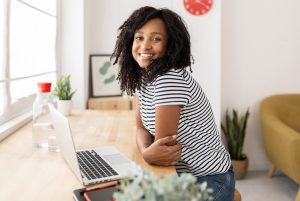 A imagem mostra uma mulher sentada, com os braços em uma mesa com um notebook e ela sorri para câmera e ilustra o texto: Visto de trabalho brasil: quais as opções e como solicitar? da Koetz Advocacia.