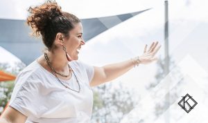 A fotografia mostra uma mulher sorrindo e acenando em um ambiente externo, e ilustra a publicação 