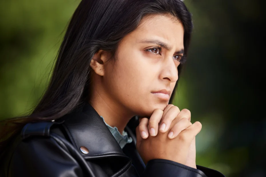 A imagem mostra uma jovem mulher, pensativa, olhando para o horizonte, com as mãos entrelaçadas, abaixo do queixo. Ilustra o texto sobre refugiado, imigrante e asilado.