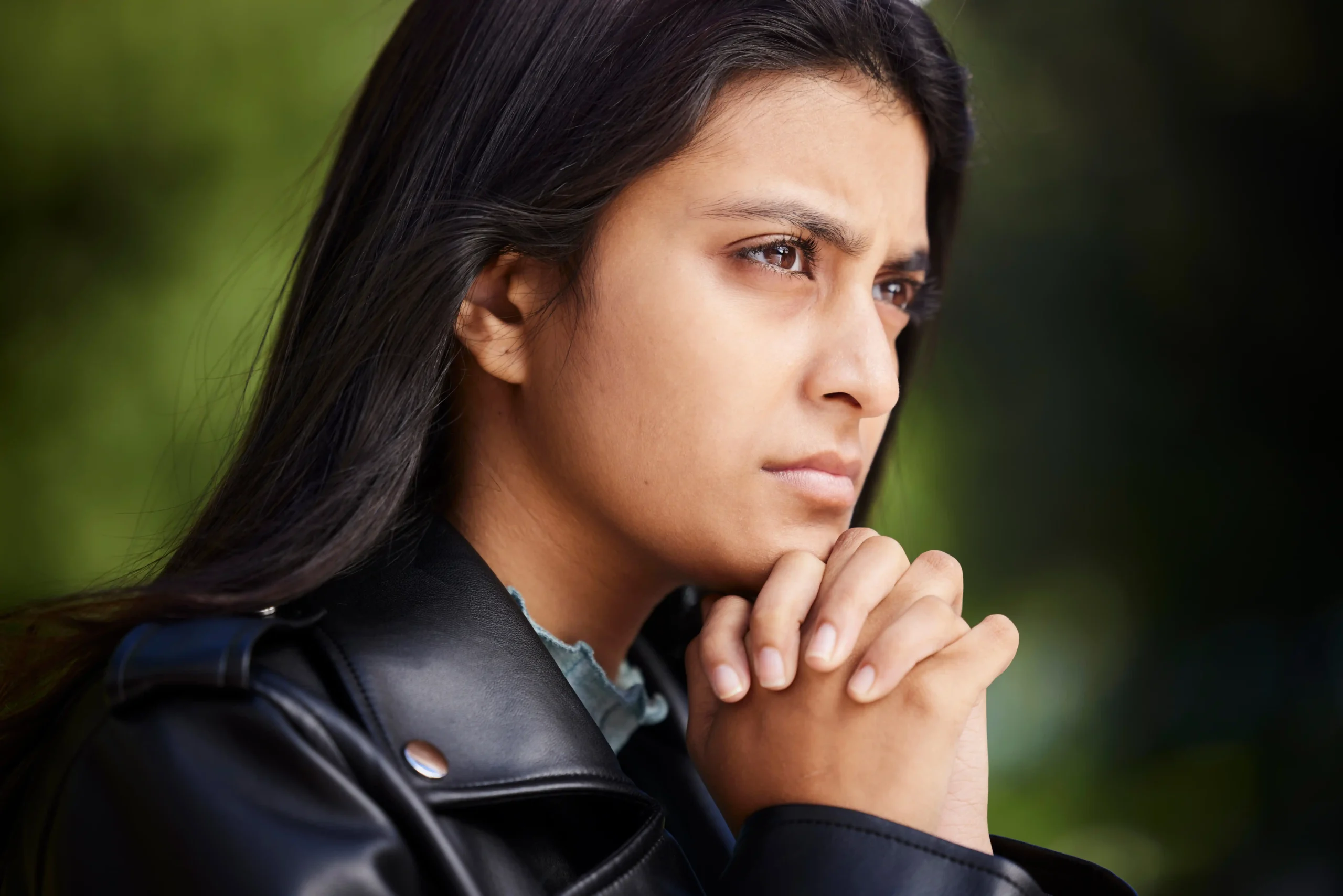 A imagem mostra uma jovem mulher, pensativa, olhando para o horizonte, com as mãos entrelaçadas, abaixo do queixo. Ilustra o texto sobre refugiado, imigrante e asilado.