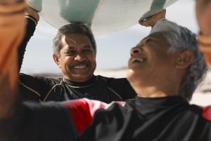 A imagem mostra um casal de idosos, segurando uma prancha de surf sobre a cabeça, sorrindo um para o outro e ilustra o texto: Los jubilados extranjeros pueden ser elegibles para Visas Brasileñas da Koetz Advocacia.