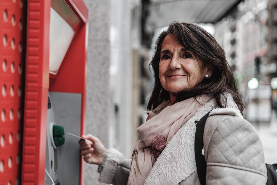A imagem mostra uma mulher adulta pegando dinheiro de uma caixa eletrônico na rua. A imagem ilustra o texto "Jubilación de inmigrantes que han trabajado en dos países o más" de Koetz Advocacia