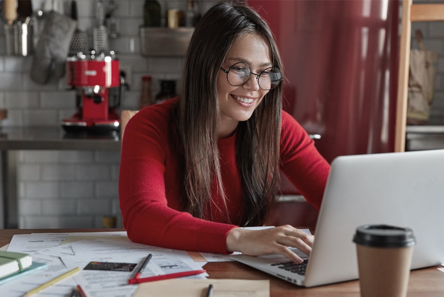 A imagem mostra uma menina jovem sorrindo, olhando para tela do seu notebook, com vários papeis a sua volta em uma mesa. A imagem ilustra o texto "Tarjeta de inmigración vencida en Brasil: multa, bloqueo y cómo renovar." de Koetz Advocacia