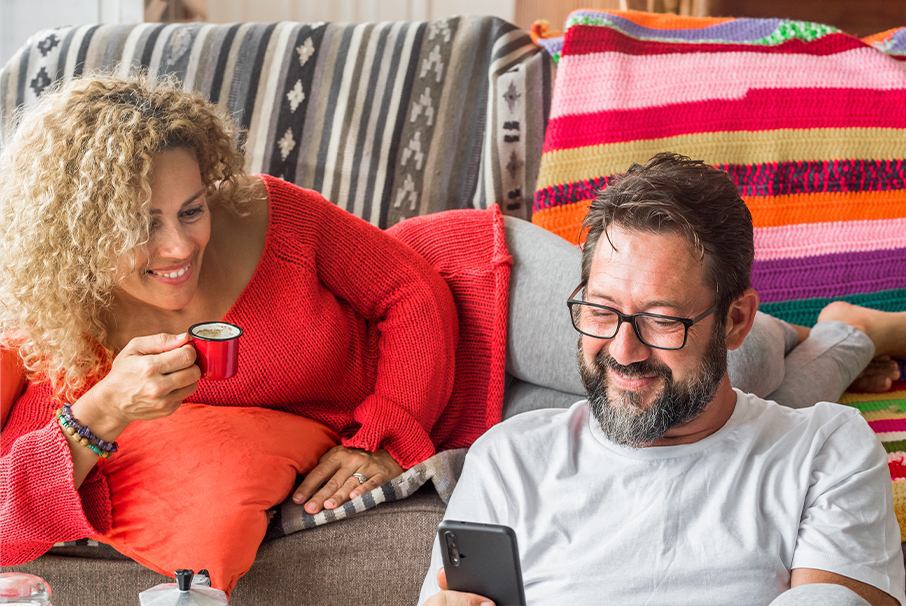 A imagem mostra um casal sorrindo, enquanto ela está no sofá com uma xícara de café e ele no chão, olhando para o celular e ilustra o texto: Modalidades de autorización de residencia en Brasil da Koetz Advocacia.