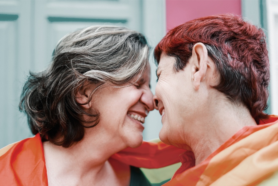 A imagem mostra um casal de lésbicas idosas vestindo a bandeira de arco-íris LGBT. A imagem ilustra o texto "Visa de unión estable en Brasil para extranjeros: ¿cómo obtenerla?" de Koetz Advocacia
