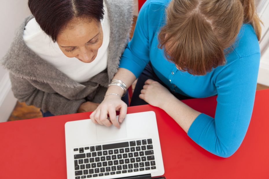 A imagem mostra uma visão de cima, onde é possível encontrar duas mulheres verificando um notebook em uma mesa e ilustra o texto: Divórcio on-line: como funciona? da Koetz Advocacia.