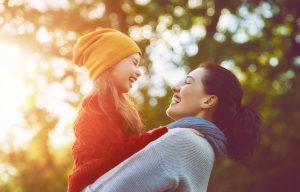 A imagem mostra uma mãe carregando sua filha, enquanto as duas sorriem em um ambiente externo e ilustra o texto: Cidadania brasileira: Qual modo mais rápido de obter o passaporte? da Koetz Advocacia.