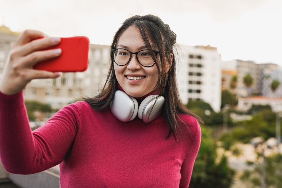 A imagem mostra uma mulher sorrindo, enquanto posa para uma selfie e usa fones de ouvido, ao ar livre e ilustra o texto: Como Revalidar e Reconhecer Diplomas Estrangeiros no Brasil da Koetz Advocacia.