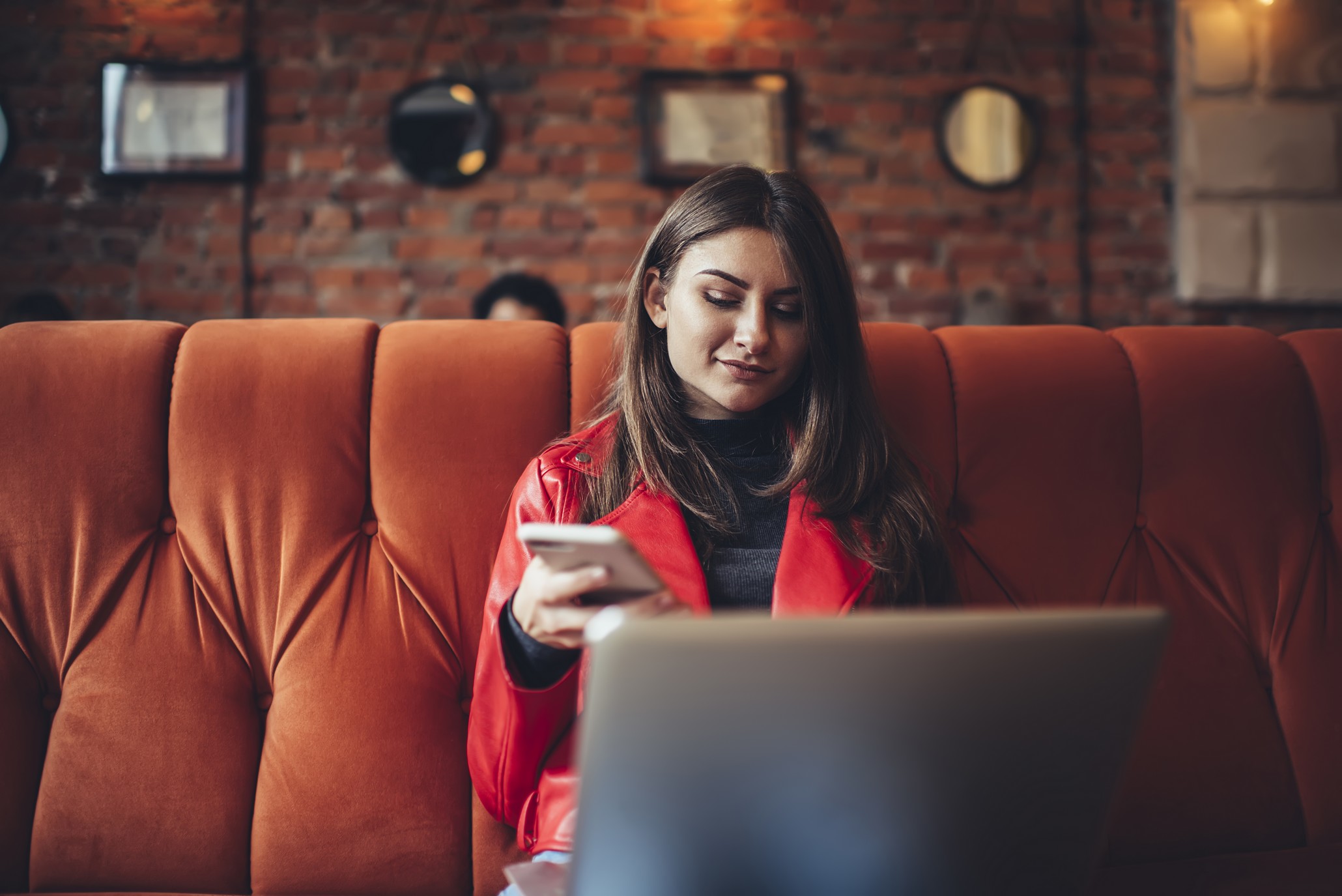 A imagem mostra uma mulher sorrindo, enquanto está sentada em um sofá, segura um celular em uma das mãos e usa o notebook em outra e ilustra o texto: How to Revalidate and Recognize Foreign Diplomas in Brazil da Koetz Advocacia.