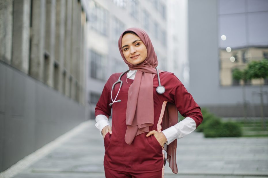 A imagem mostra uma médica em pé, usando roupas ciirúrgias, hijab e olhando para câmera, enquanto sorri e ilustra o texto: How to revalidate medical diplomas da Koetz Advocacia.