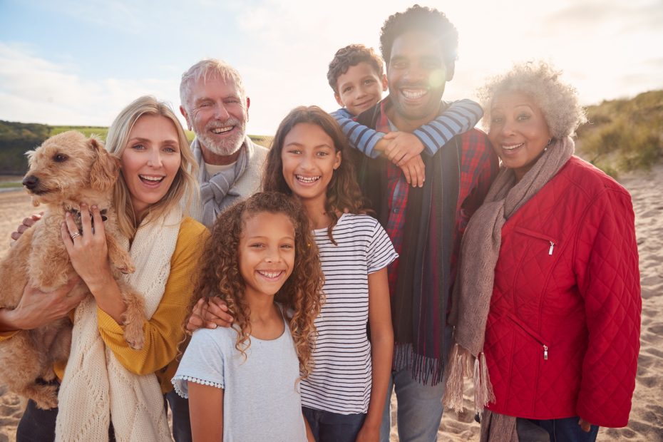 A imagem mostra uma família reunida (composta por adultos, jovens, crianças e idosos) em uma área ao ar live, em uma praia ensolarada e ilustra o texto: Vantagens da Sucessão Familiar no Brasil – comparativo com os EUA da Koetz Advocacia. bem estar social