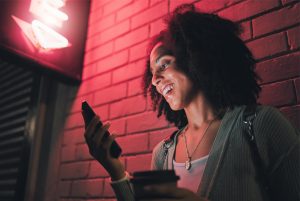 La imagen muestra a una mujer sonriente apoyada contra una pared mientras mira su teléfono celular. ilustrando el articulo 