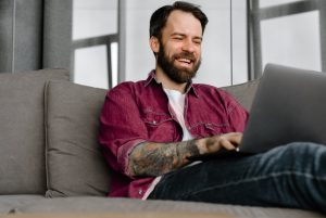 The image shows a smiling man with his laptop while sitting on the sofa. ilustrating the article 