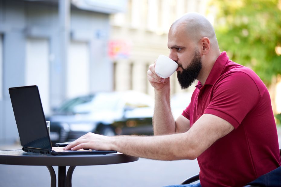 A imagem mostra um empresário trabalhando em um laptop, enquanto toma uma xícara de café ao ar livre e ilustra o texto: Can I have investments in Brazil if I live abroad? da Koetz Advocacia.