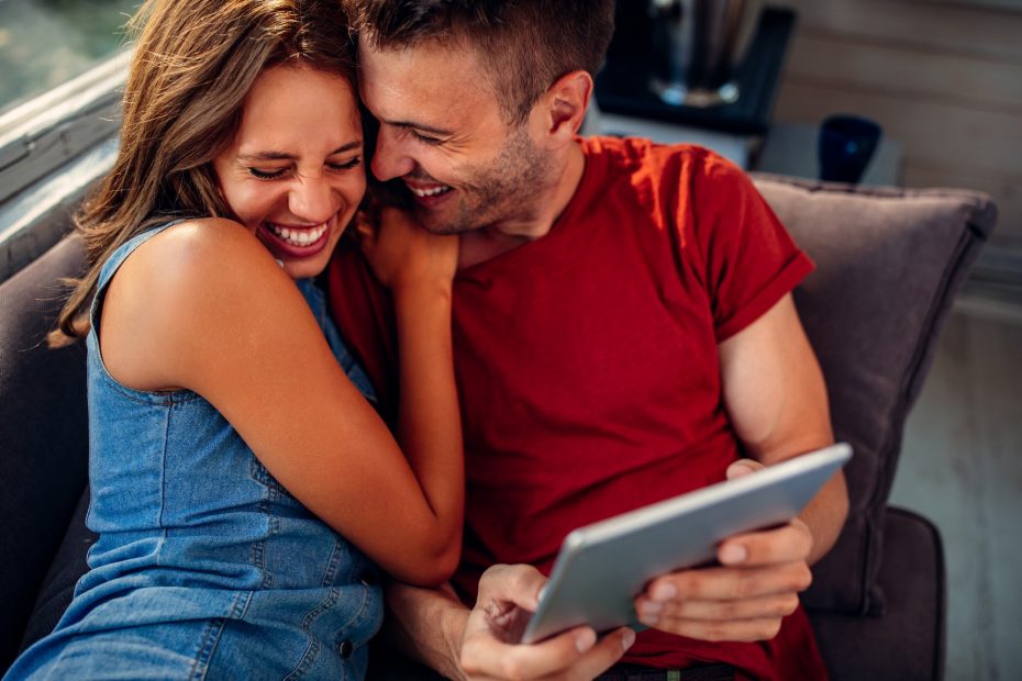 A imagem mostra um casal sorrindo, abraçados, enquanto estão sentados em um sofá e checando um tablet que está nas mãos do homem e ilustra o texto: Holding Familiar: o que é, vantagens e como abrir a sua.