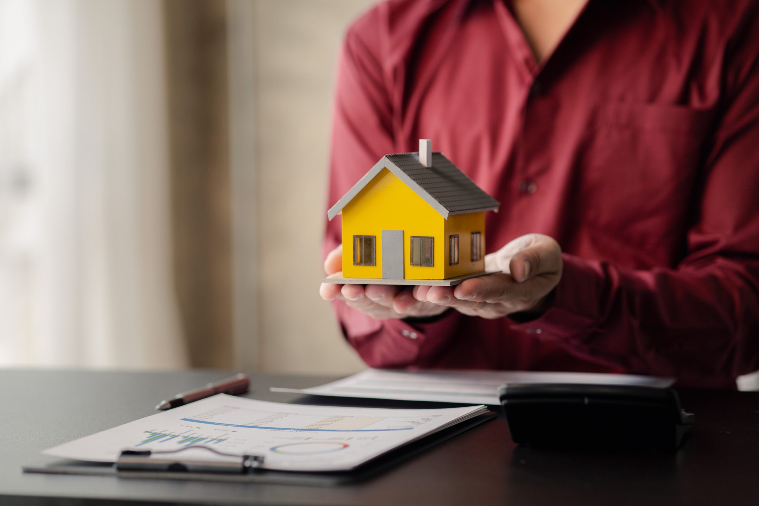 A imagem mostra um homem segurando uma casa em miniatura nas mãos, junto de documentos com gráficos, e ilustra o texto: Holding patrimonial: o que é e quais as vantagens? da Koetz Advocacia.