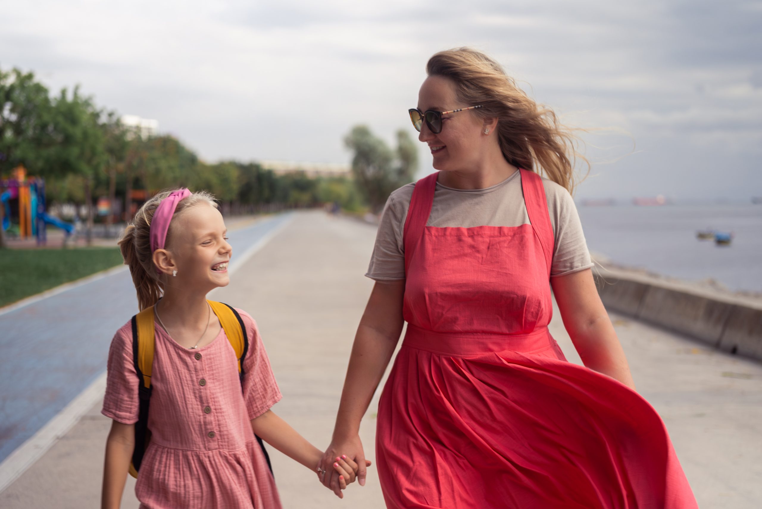 A imagem mostra uma mãe, de mãos dadas com sua filha criança, que está usando uma mochila nas costas e ambas sorriem e ilustra o texto: Transferindo os Filhos para a escola em Portugal: o que preciso saber? da Koetz Advocacia.