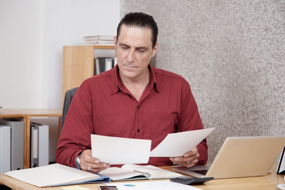 A imagem mostra um homem de camisa social analisando documentos em sua mesa de trabalho, e ilustra a publicação "Documentos necessários para fazer testamento em vida", da Koetz Advocacia.