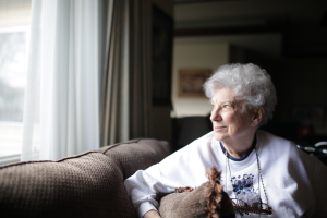 The image shows an elderly woman, sitting on a sofa, holding a cushion, while looking at the horizon through a window and illustrates the text: Brazilian pension for a foreign widow or widower by Koetz Advocacia.