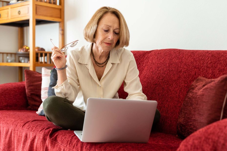 A imagem mostra uma mulher na melhor idade usando o computador no sofá, e ilustra a publicação "Quanto custa para fazer um testamento em vida?", da Koetz Advocacia.