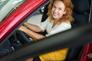 A imagem mostra uma mulher sorrindo, dentro de um carro, no banco do motorista, com as mãos no volante e ilustra o texto: Importação e exportação de veículos próprios da Koetz Advocacia.
