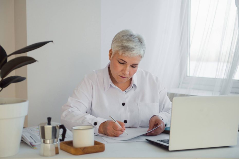 A imagem mostra uma mulher da terceira idade lendo documentos em frente ao computador, e ilustra a publicação da Koetz Advocacia: Testamento cerrado: o que é e quando usar?