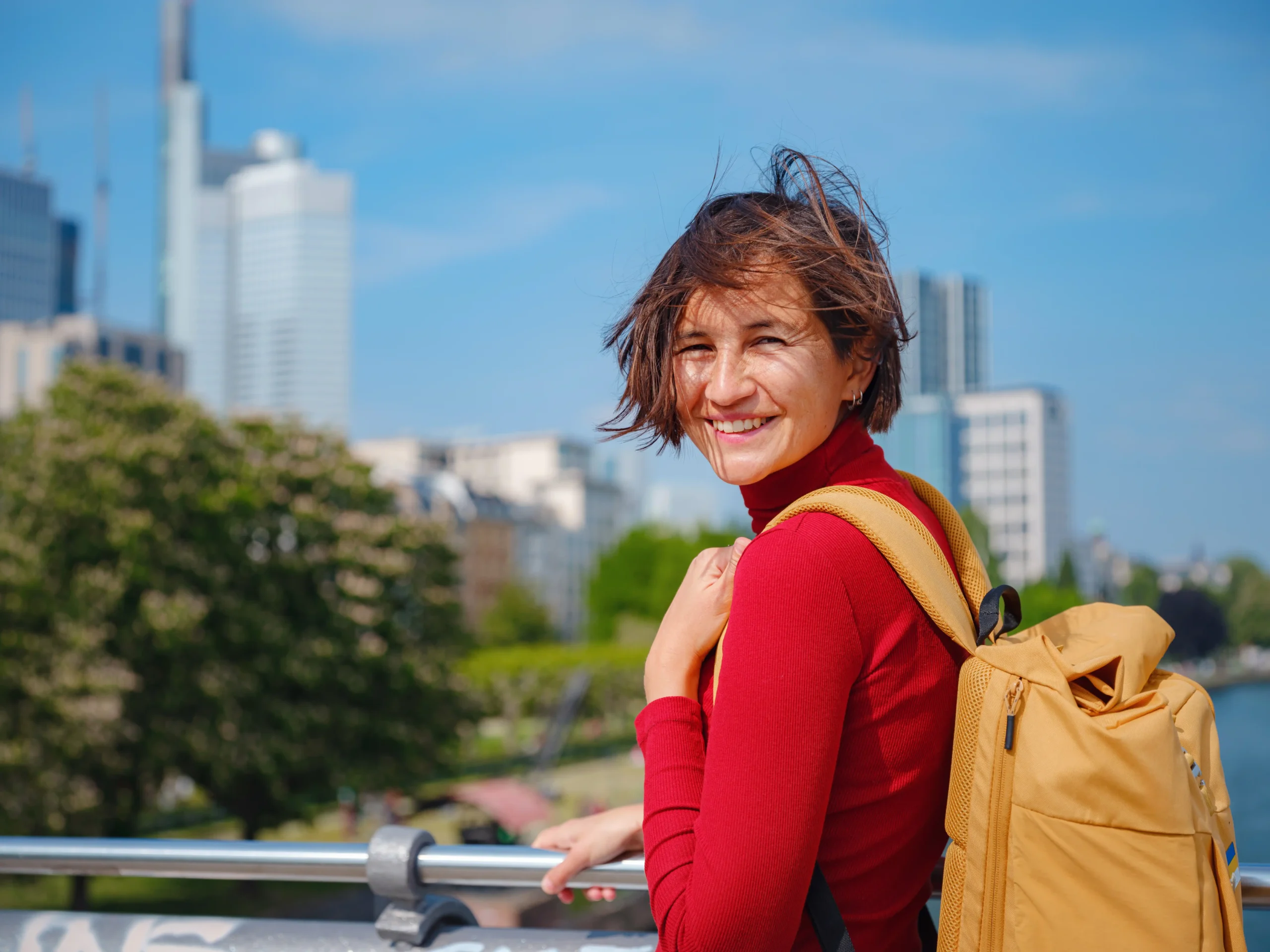 A imagem mostra uma mulher sorridente, ao ar livre, em uma grande cidade, com mochilas nas costas. Ela olha para a câmera e ilustra o texto sobre imigrante e seus direitos.