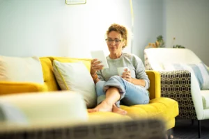 La imagen muestra a una mujer de mediana edad leyendo un ebook en una tableta, sentada cómodamente en el sofá de su casa. Ilustra el texto sobre propiedades en Brasil.