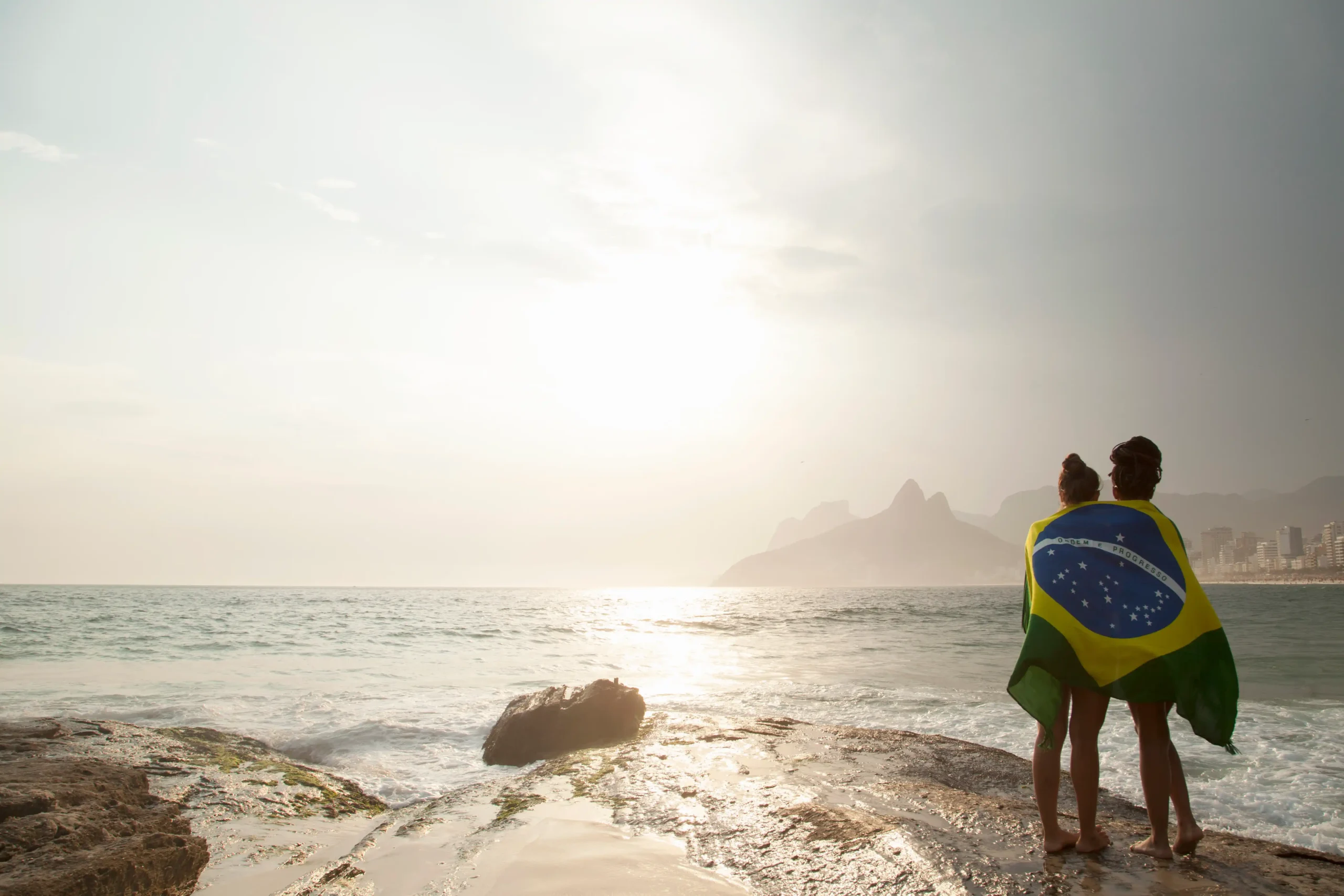 A imagem mostra a vista traseira de duas jovens embrulhadas na bandeira brasileira na praia de Ipanema, Rio de Janeiro, Brasil. E ilustra o texto sobre imigração para o Brasil.
