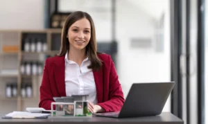 La imagen muestra a una mujer joven sonriendo a la cámara, trabajando con una computadora portátil en el escritorio de la oficina y la pequeña casa de al lado. Y ilustra las ventajas de Holding Patrimonial.