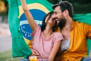 La imagen muestra a una pareja joven, sonriente, sosteniendo la bandera brasileña, al aire libre. Ilustra el texto sobre la inmigración a Brasil.