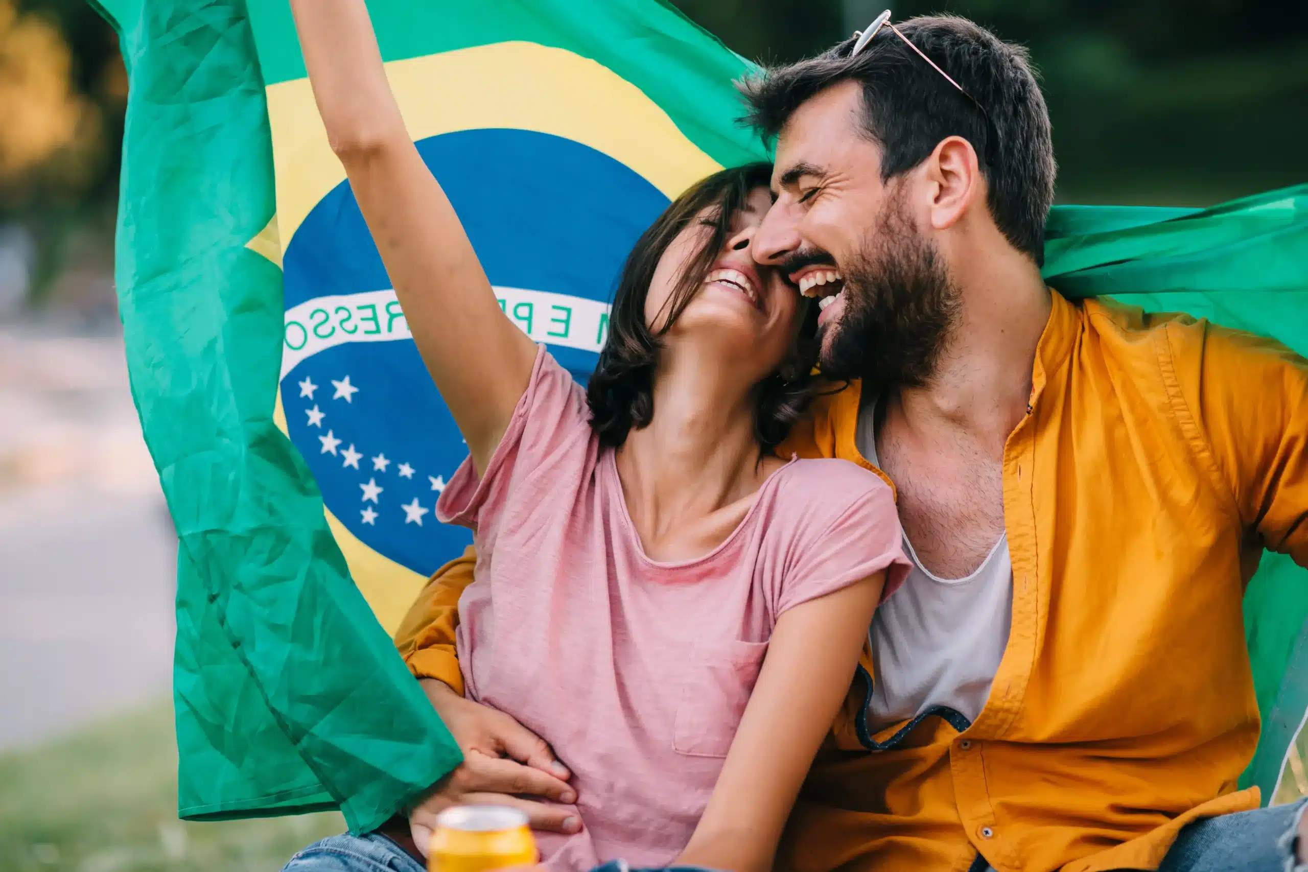 La imagen muestra a una pareja joven, sonriente, sosteniendo la bandera brasileña, al aire libre. Ilustra el texto sobre la inmigración a Brasil.