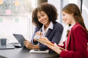 La imagen muestra dos mujeres vestidas formalmente, en un ambiente corporativo, conversando y sonriendo e ilustra el texto: Cómo obtener una visa brasileña invirtiendo en innovación de Koetz Advocacia.