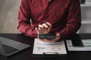 La imagen muestra unas manos masculinas resaltadas, utilizando una calculadora, cerca de documentos financieros e ilustra el texto: PL 4173 y la Nueva Tributación de las Sociedades Offshore de Koetz Advocacia.