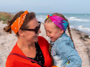 La imagen muestra a una madre y a su hija jugando en la playa, mientras la hija está en el regazo de su madre y ambas sonríen. La imagen ilustra el texto: ¿Puede un inversor extranjero en Brasil obtener una visa para toda la familia? de Koetz Advocacia.