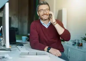 La imagen muestra a un empresario sonriente hablando por un teléfono celular mientras está sentado en una silla de oficina, apoyado en una mesa e ilustra el texto: Préstamo Convertible en Brasil: ventajas del formato en la Colección de Inversión Extranjera de Koetz Advocacia.