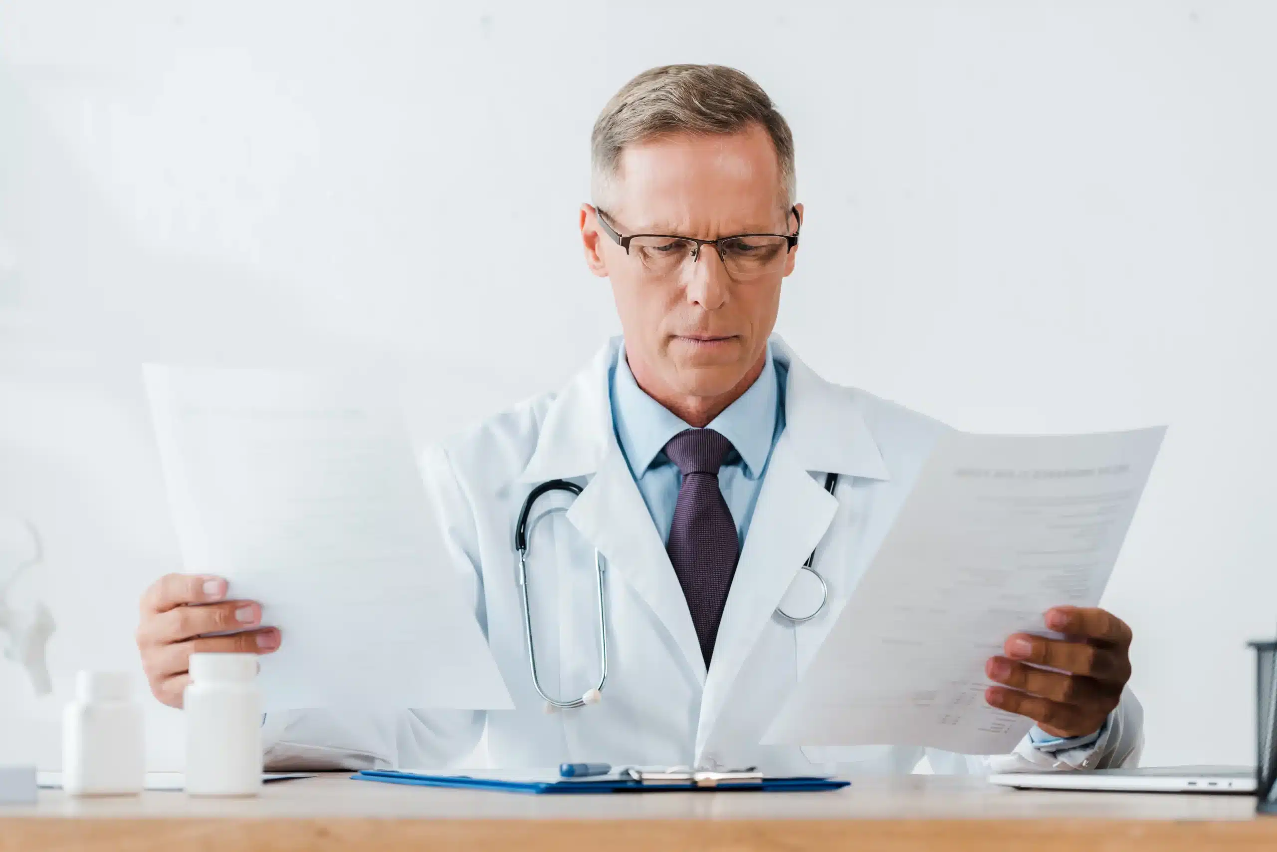 A imagem mostra um médico de meia idade, com jaleco e estetoscópio em volta do pescoço, analisando documentos. Ilustra o texto sobre prontuário médico.
