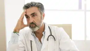 A imagem mostra um médico em seu escritório, de jaleco e estetoscópio em volta do pescoço, segurando a cabeça com uma das mãos, de forma preocupada. Ilustra o texto sobre difamação médica.
