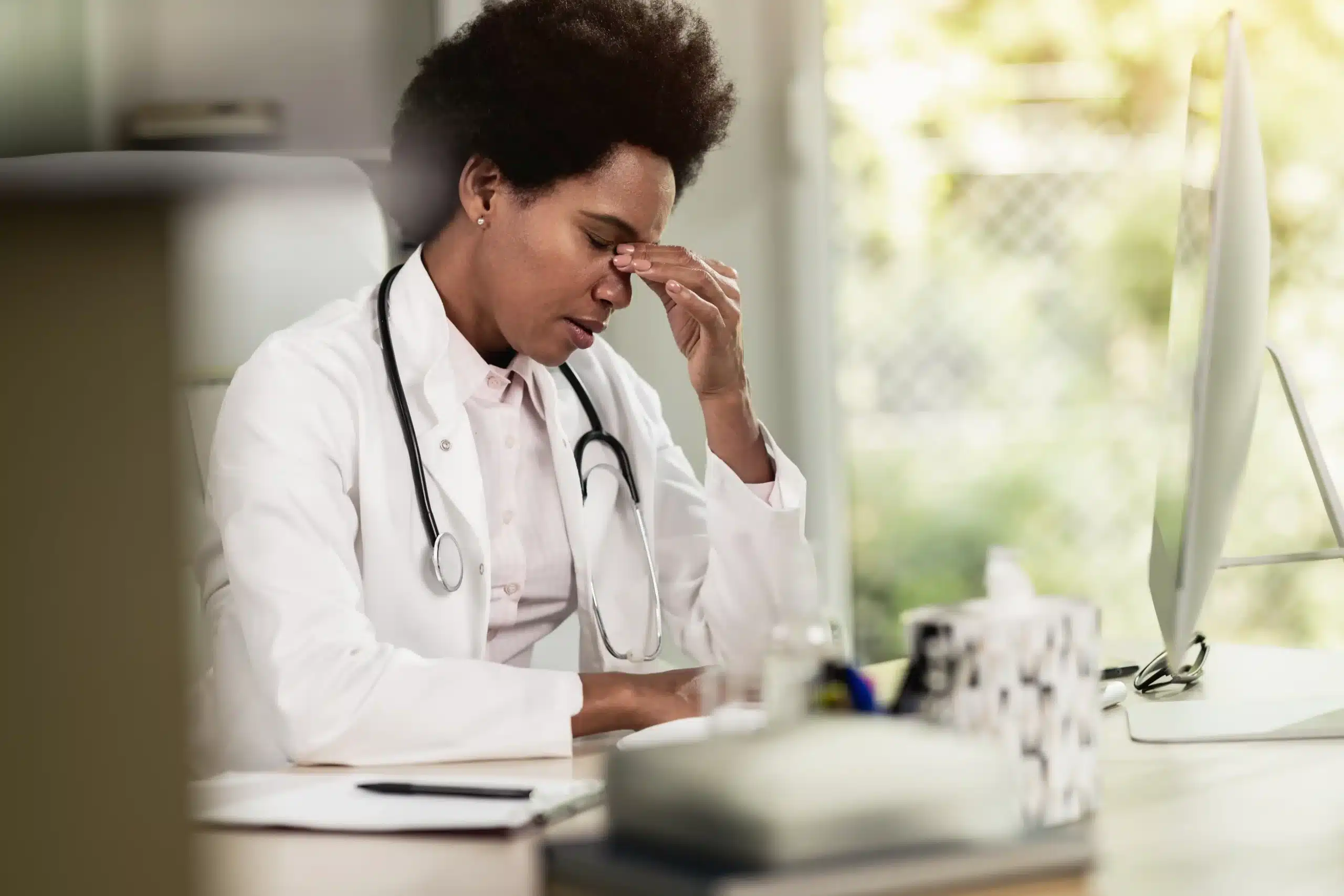 A imagem mostra uma médica negra, usando jaleco e estetoscópio em volta do pescoço, sentada em uma cadeira, em seu consultório, com uma expressão preocupada. Ilustra o texto sobre médico pode ser processado civil e criminalmente.