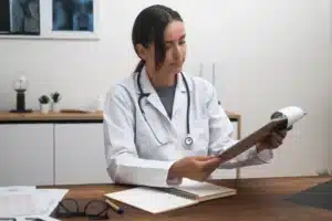 A imagem mostra uma médica estudando documentos em prancheta sentada à mesa em consultório iluminado. Ilustra o texto sobre coworking médico.
