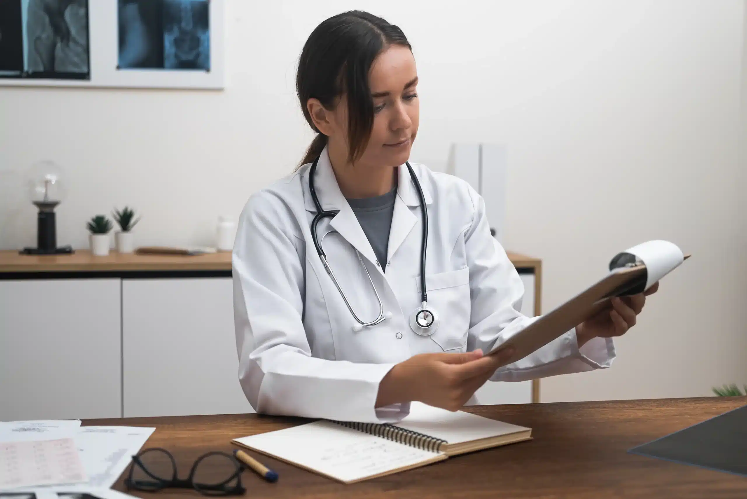A imagem mostra uma médica estudando documentos em prancheta sentada à mesa em consultório iluminado. Ilustra o texto sobre coworking médico.