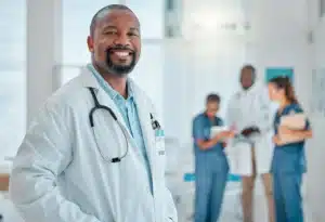 A imagem mostra um médico, sorrindo para a câmera, em ambiente hospitalar. Ele usa jaleco e estetoscópio em volta do pescoço. Ilustra o texto sobre contrato para médico plantonista.