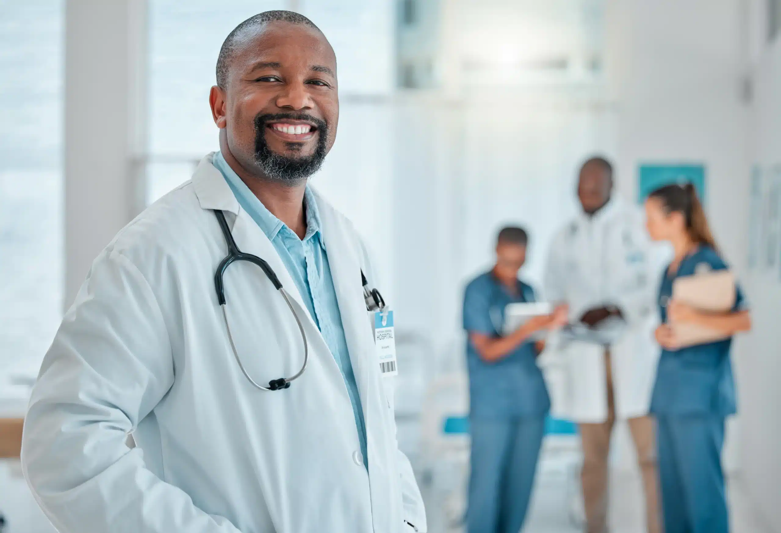 A imagem mostra um médico, sorrindo para a câmera, em ambiente hospitalar. Ele usa jaleco e estetoscópio em volta do pescoço. Ilustra o texto sobre contrato para médico plantonista.