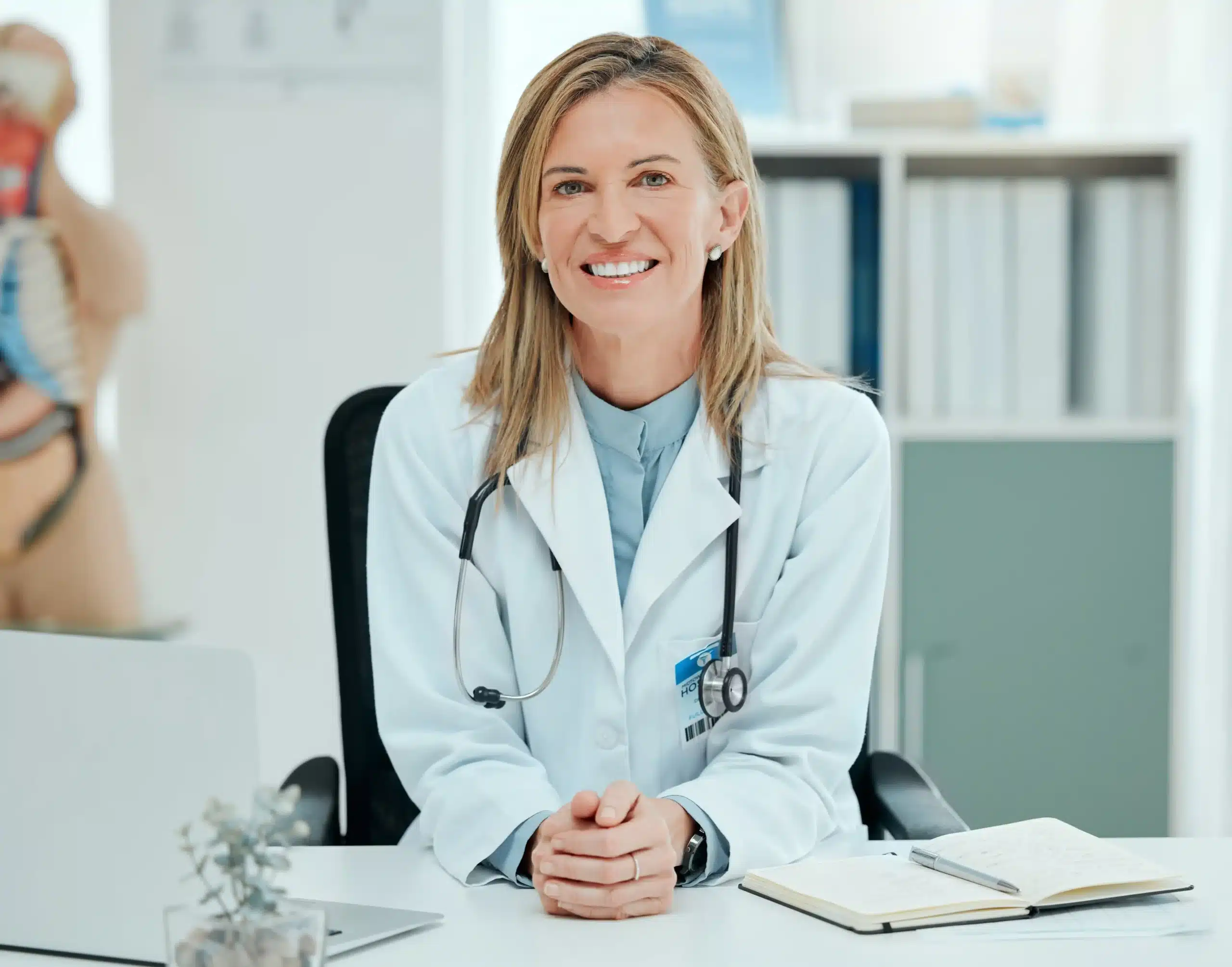 A imagem mostra uma médica de meia idade, com cabelos loiros, em seu consultório. Ela usa jaleco, estetoscópio em volta do pescoço e sorri para a câmera. Ilustra o texto sobre responsabilidade civil do médico.