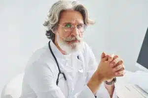 A imagem mostra um médico sênior, com expressão sérIa, em seu consultório. Ele olha para a câmera e veste jaleco e estetoscópio em volta do pescoço. Ilustra o texto sobre seguro erro médico.